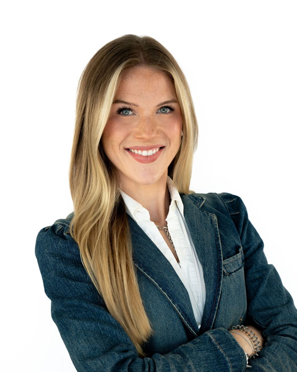 Smiling blonde woman in denim blazer and white shirt with arms crossed, professional headshot on white background.