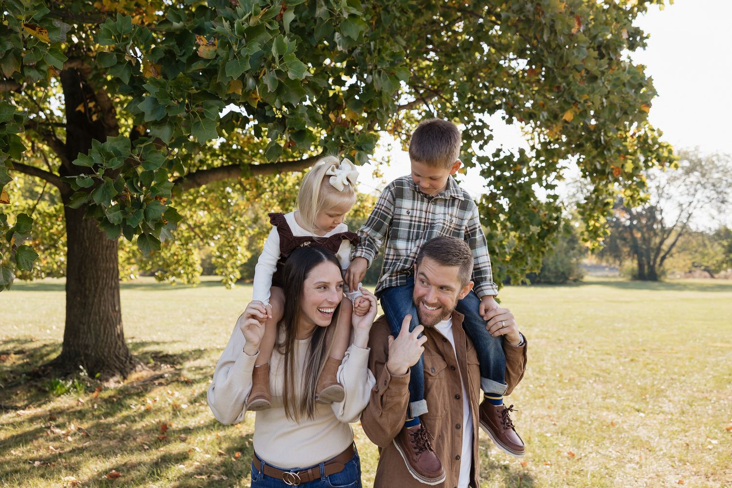 mom and dad each holding a child on their shoulders while everyone smiles and plays together under a big tree in overland park kansas