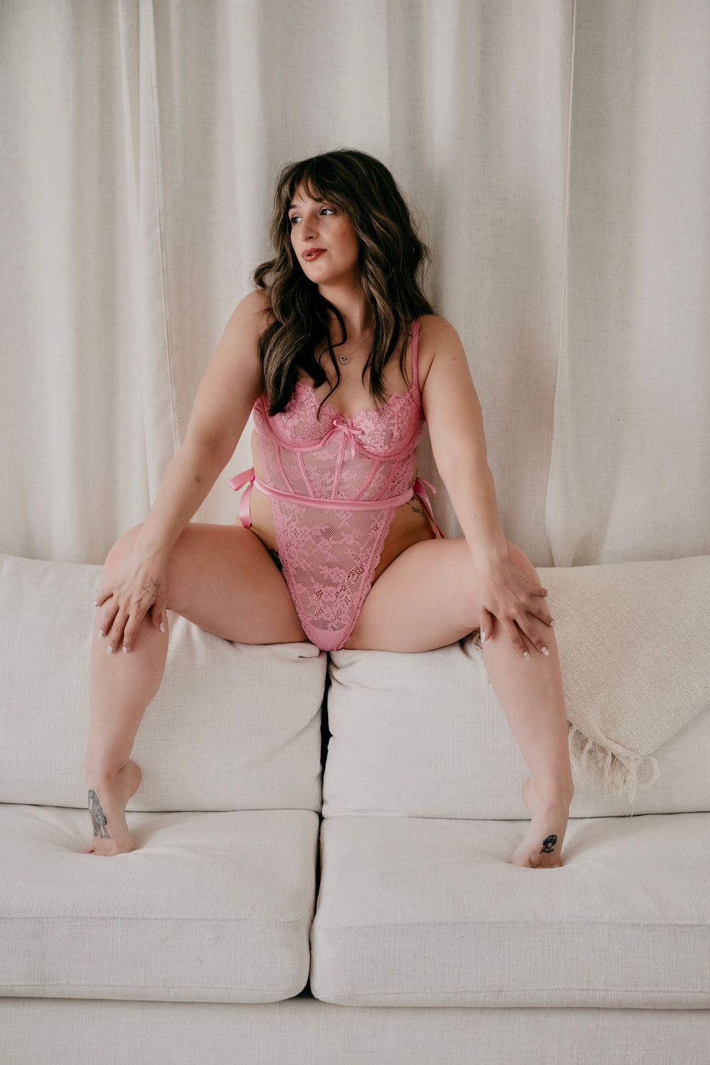 Woman in pink lace bodysuit posing on white sofa with curtain backdrop.