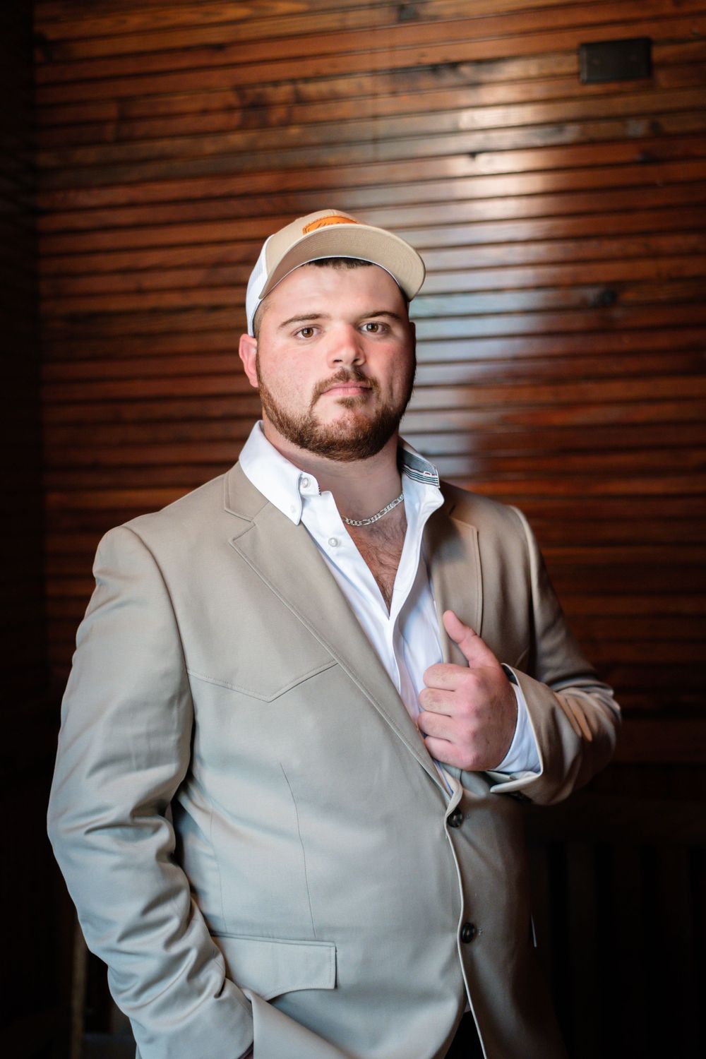 Portrait of someone in a beige blazer and baseball cap against wooden paneled walls.