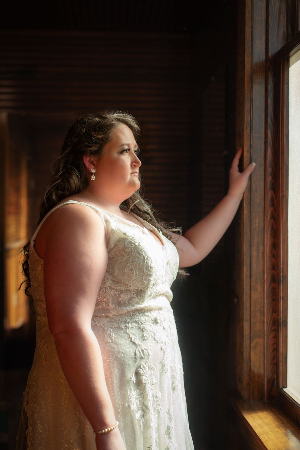 A bride in lace dress stands by window in rustic wooden interior.