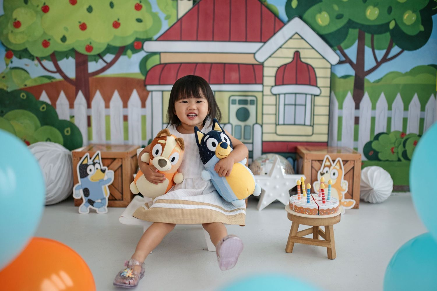 Young girl posing with Bluey character plush toys at a colorful birthday party backdrop with balloons.