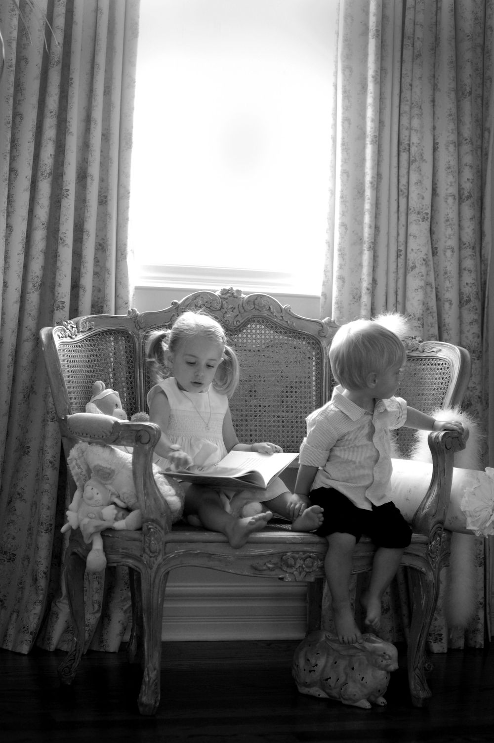 Vintage black and white photo of children reading on an ornate sofa by window drapes.