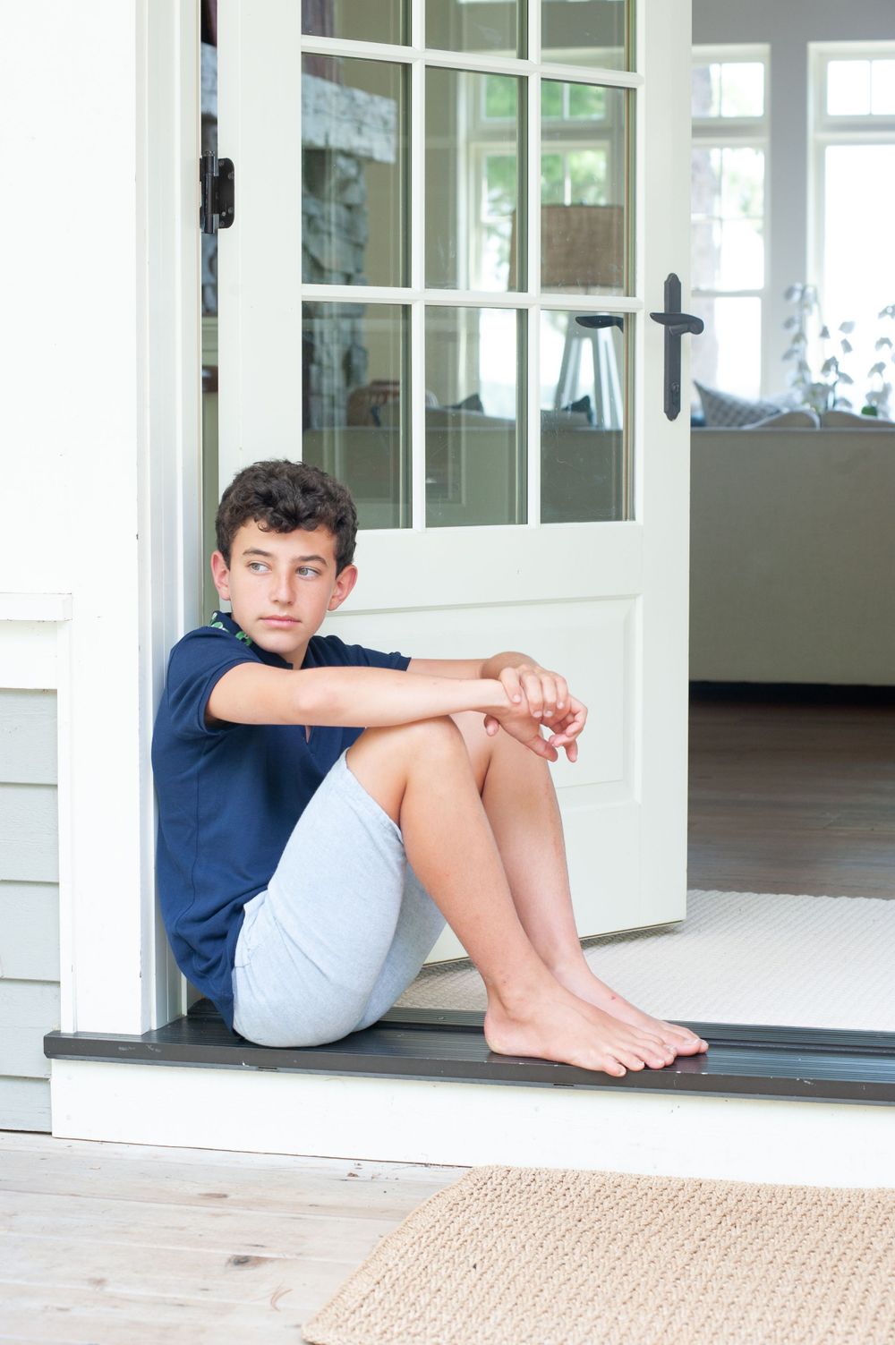 Person sitting in doorway of white house wearing navy shirt and grey shorts.