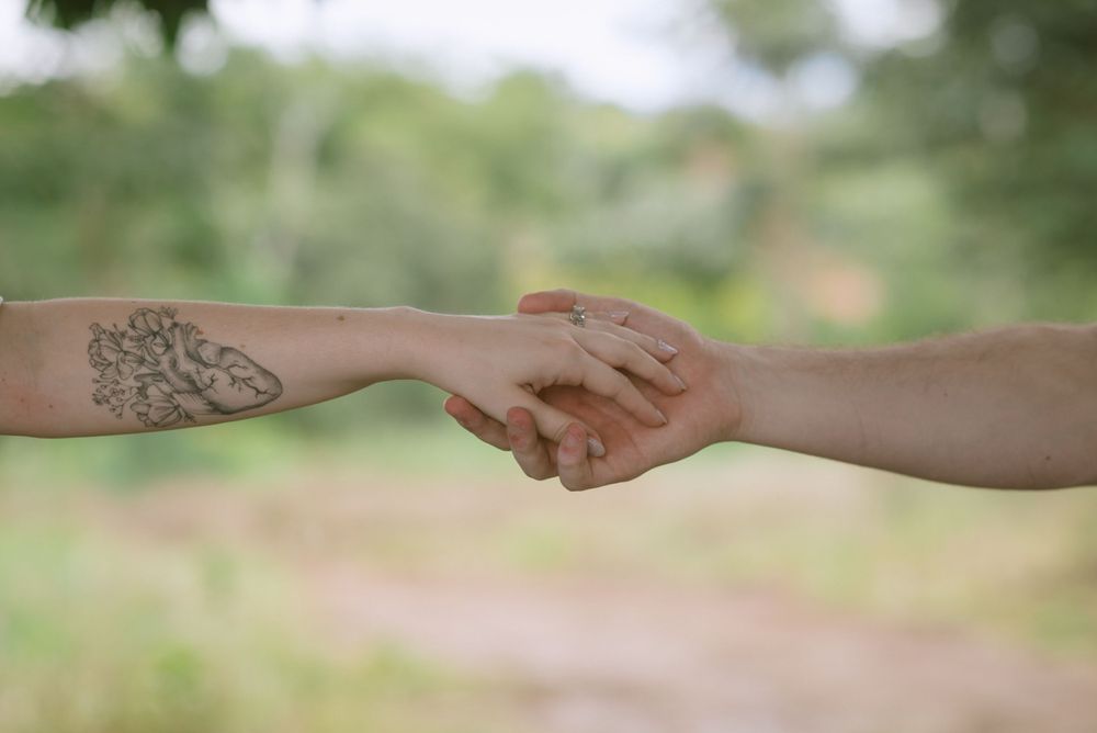 Two arms reaching out to hold hands, with one arm displaying a decorative tattoo, against a blurred natural background.