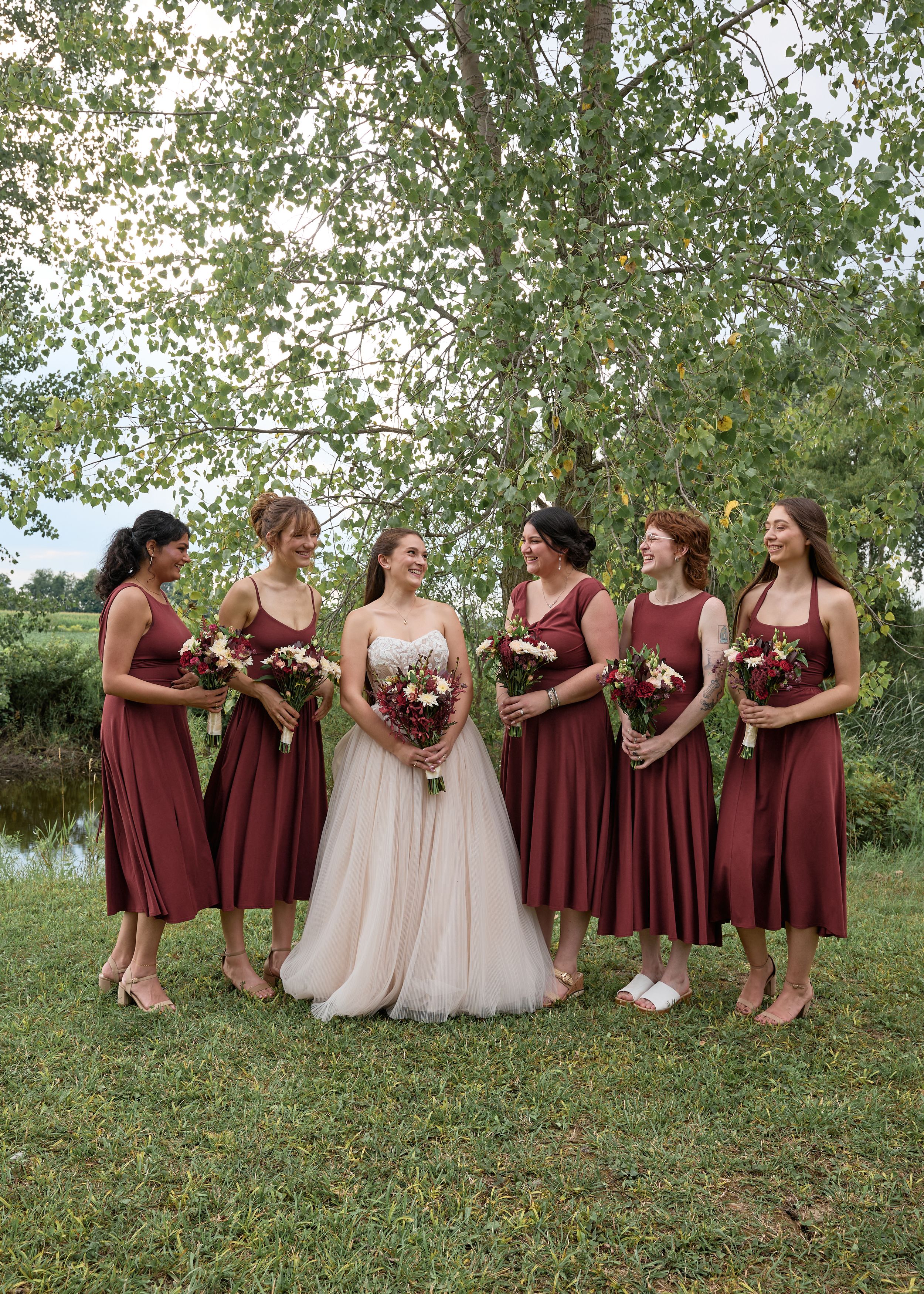 Bride in white gown with five bridesmaids in burgundy dresses holding bouquets outdoors near trees.