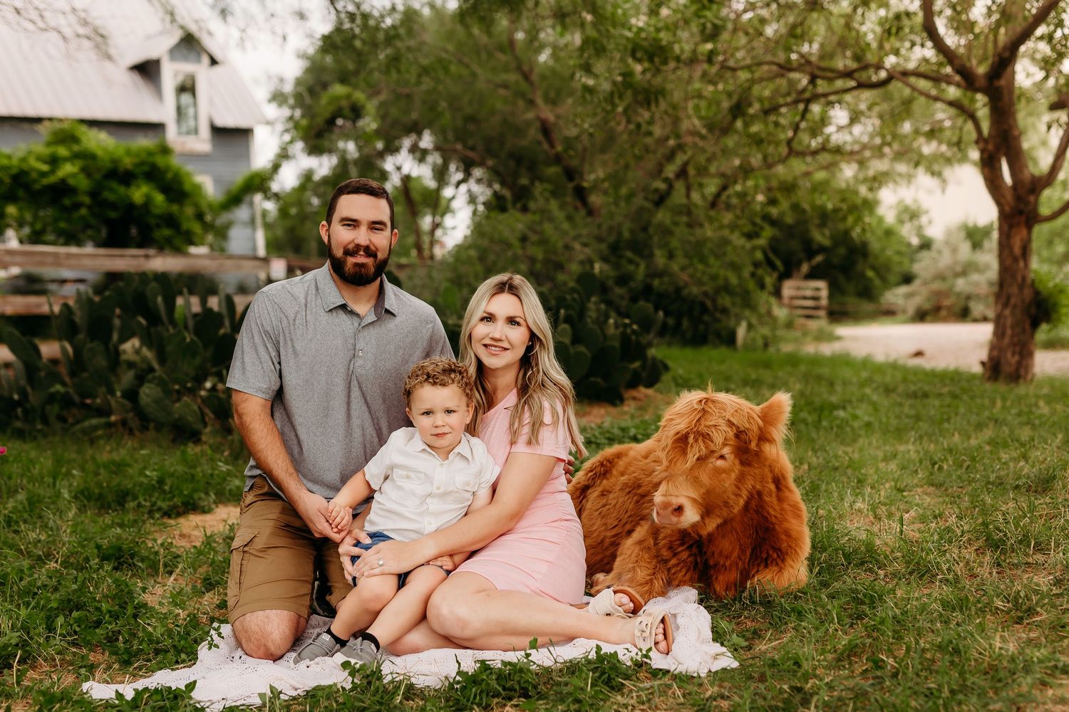 A young family sits on grass with their large golden doodle dog in a backyard garden setting.