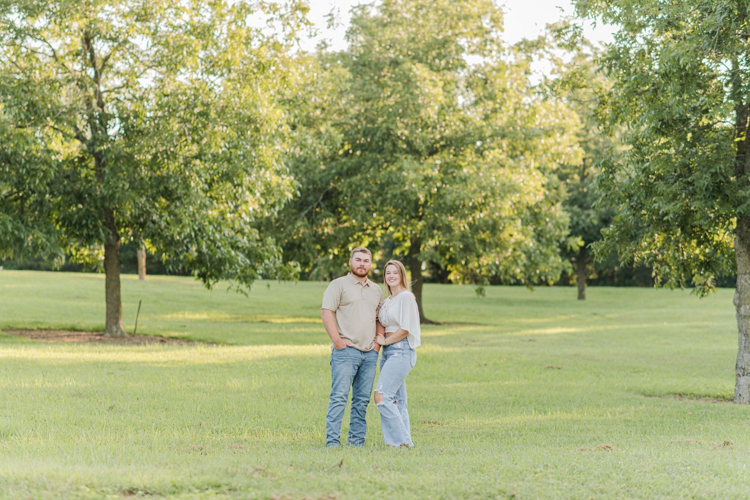 A couple stands together smiling in a lush green park surrounded by trees during golden hour.