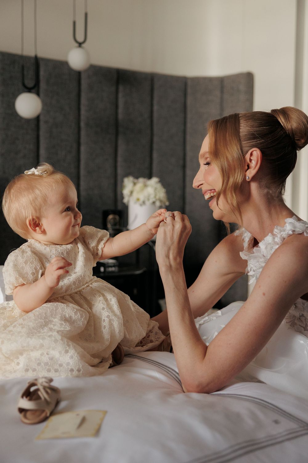 A smiling bride in a white dress sits on a bed holding hands with an adorable baby girl in a cream lace dress.