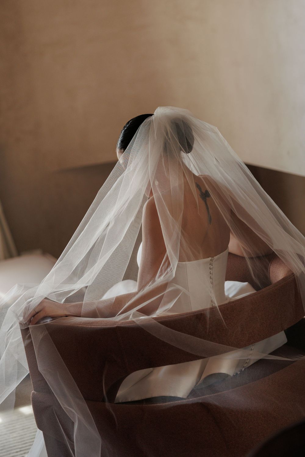 Artistic close-up photo of a sheer white wedding veil draped over a dark brown leather couch in soft natural light.