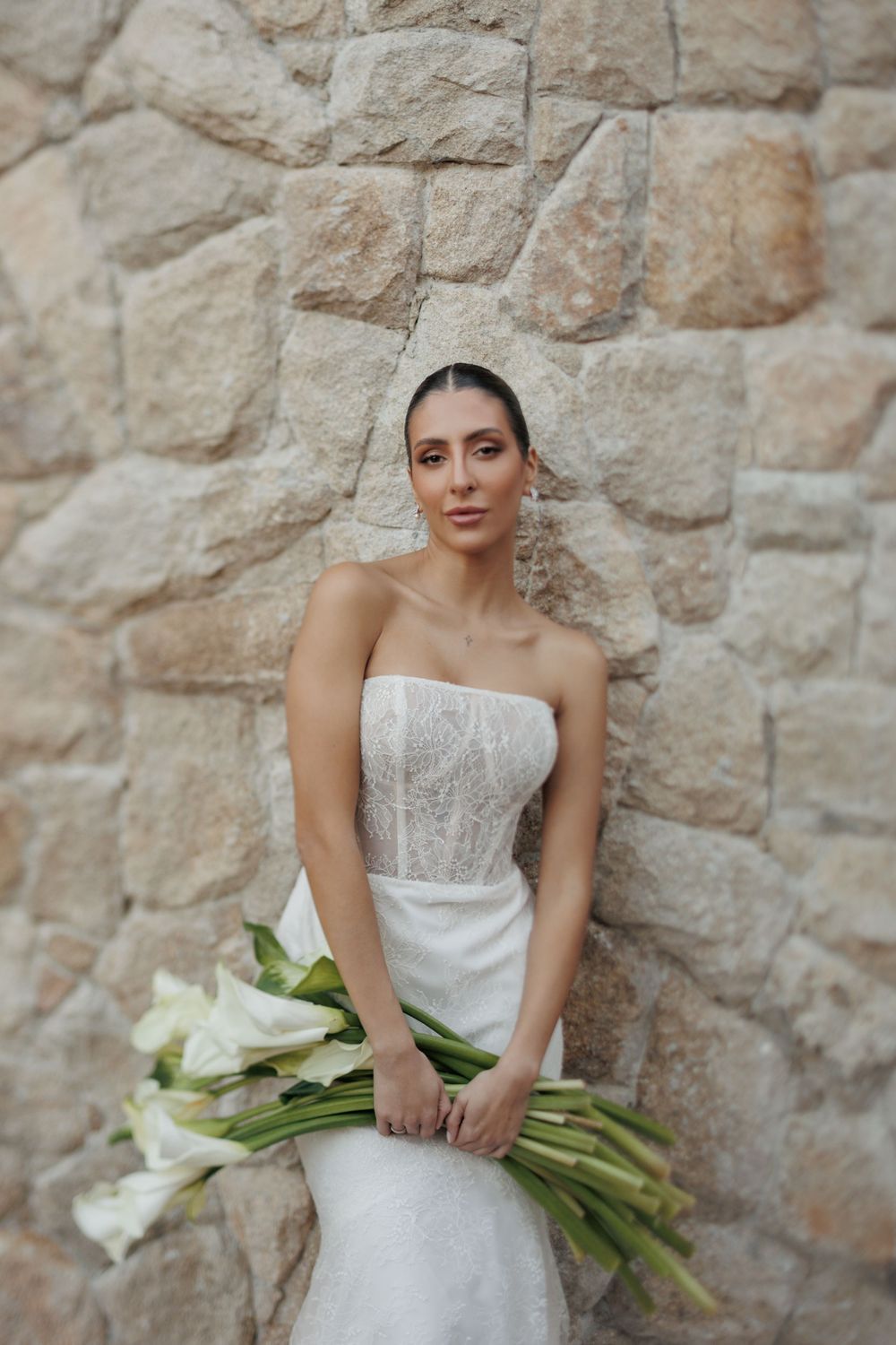 Bride in a strapless white wedding gown holds a bouquet of white calla lilies against a stone wall.