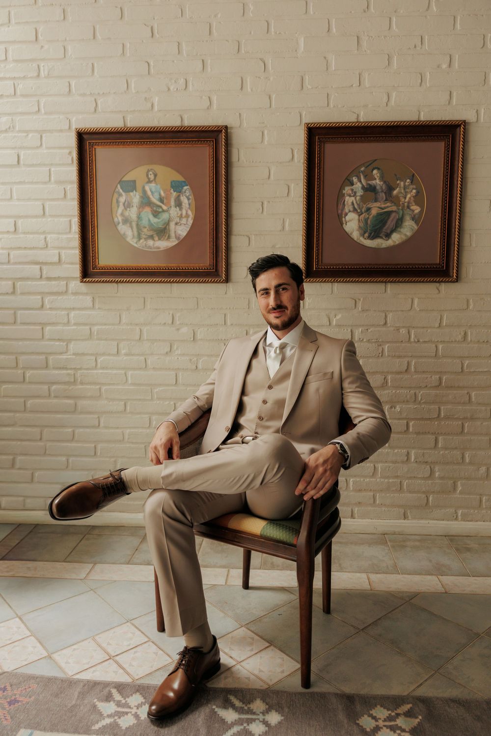 A person in a beige suit sits in different poses on a wooden chair against a white brick wall with framed artwork.