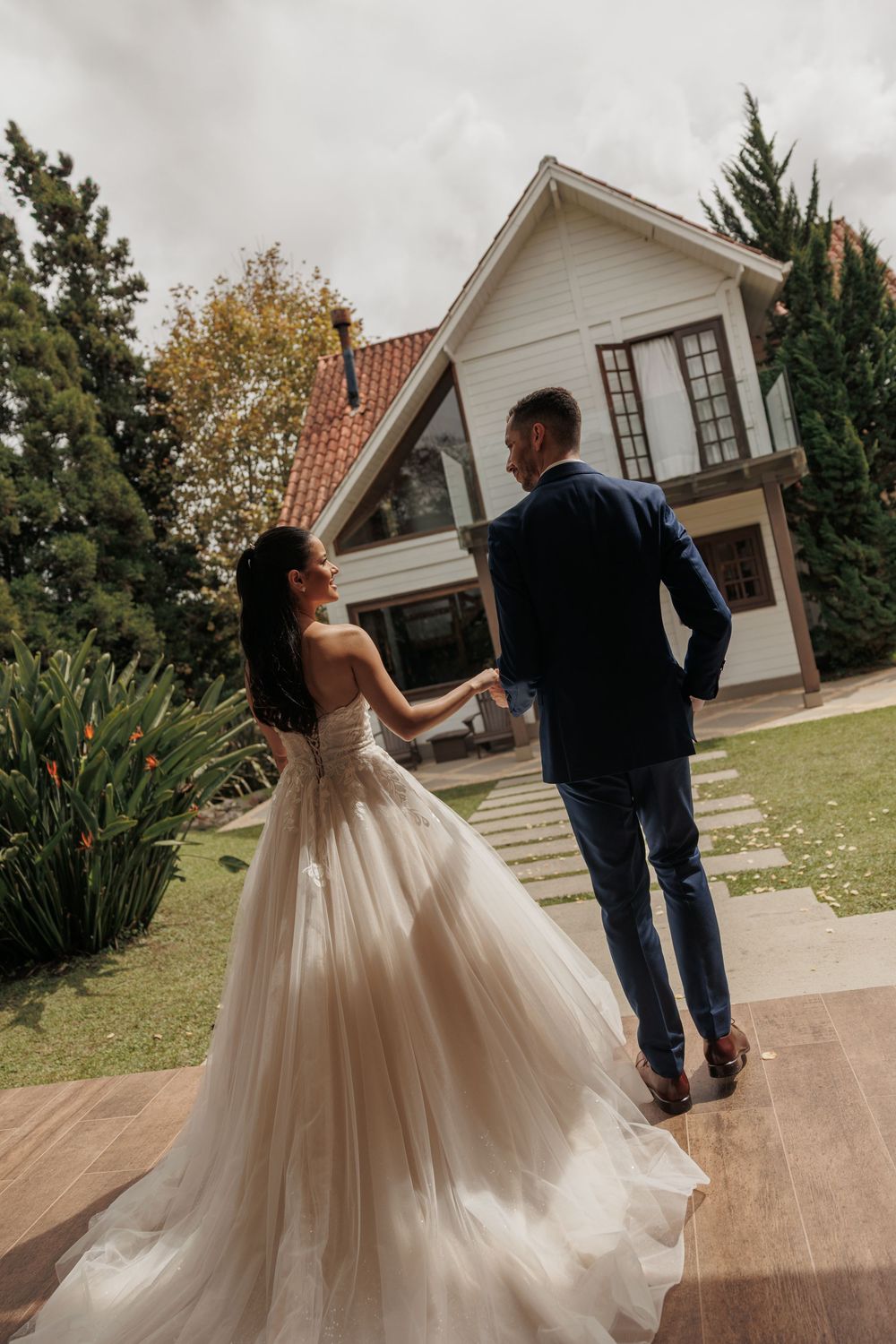 A couple in wedding attire dance together on a wooden deck with a white house and autumn trees in the background.