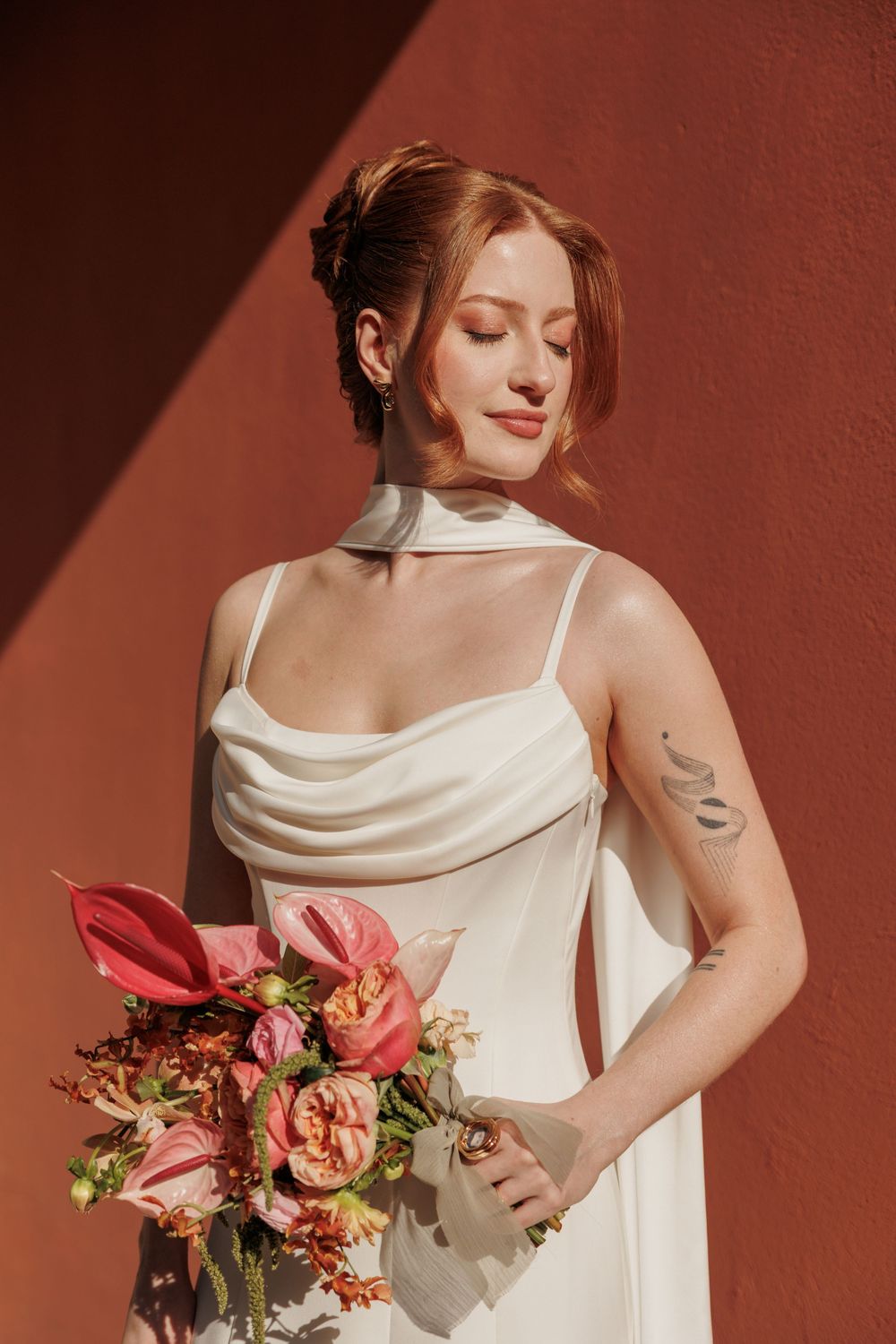 An elegant bride in a draped white gown holds a bouquet of pink flowers against a coral-colored wall.