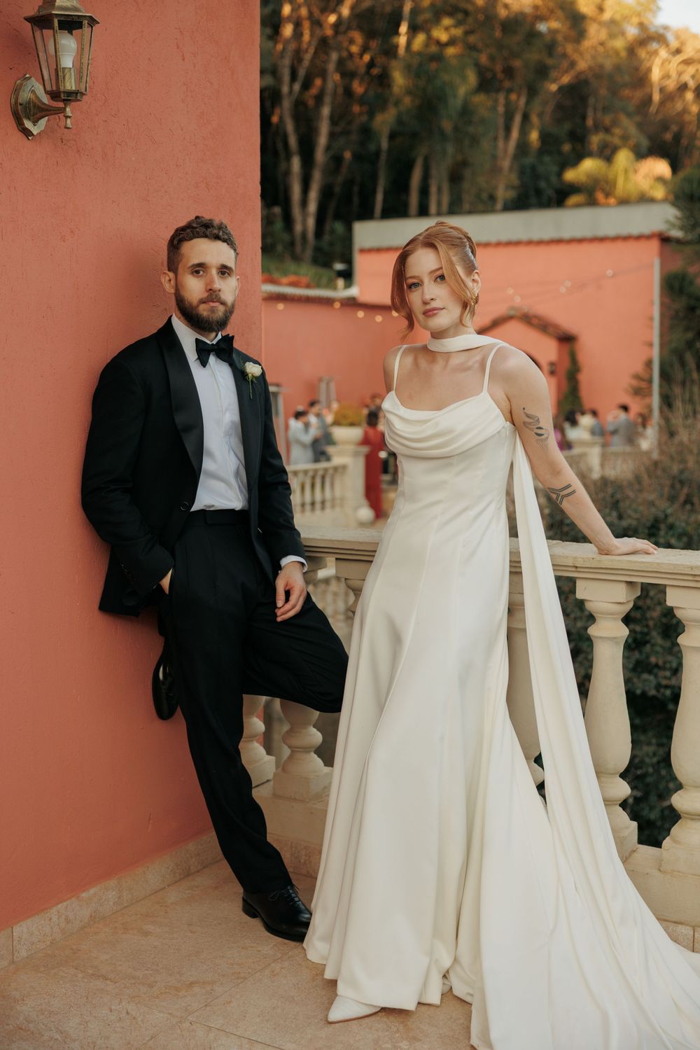 A couple in formal wedding attire poses against a coral-colored wall during golden hour at an elegant outdoor venue.