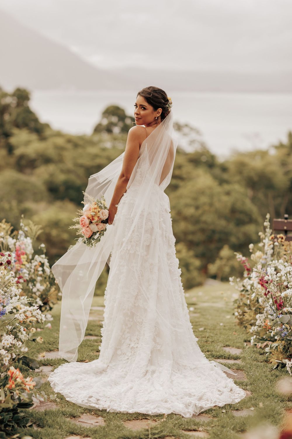 Bride in flowing lace wedding gown poses with bouquet on hillside surrounded by floral arrangements.