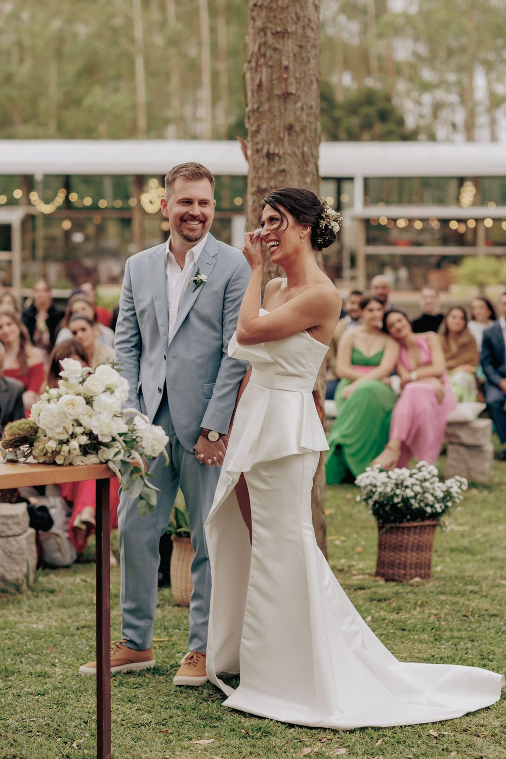 An outdoor wedding ceremony with a couple laughing at the altar surrounded by guests and floral arrangements.