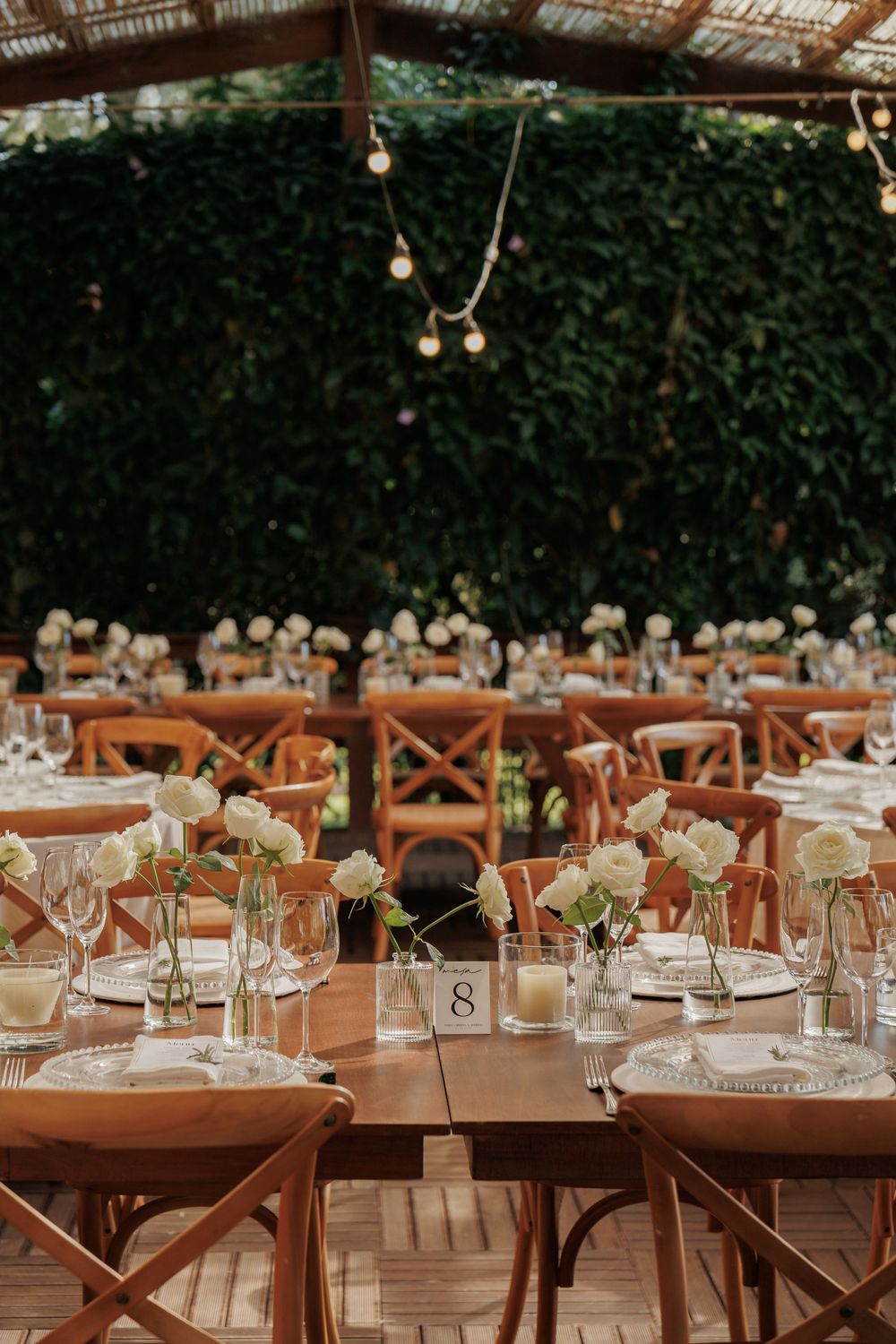 Elegant wedding reception table setup with rustic wooden chairs, white floral centerpieces, and hanging string lights.