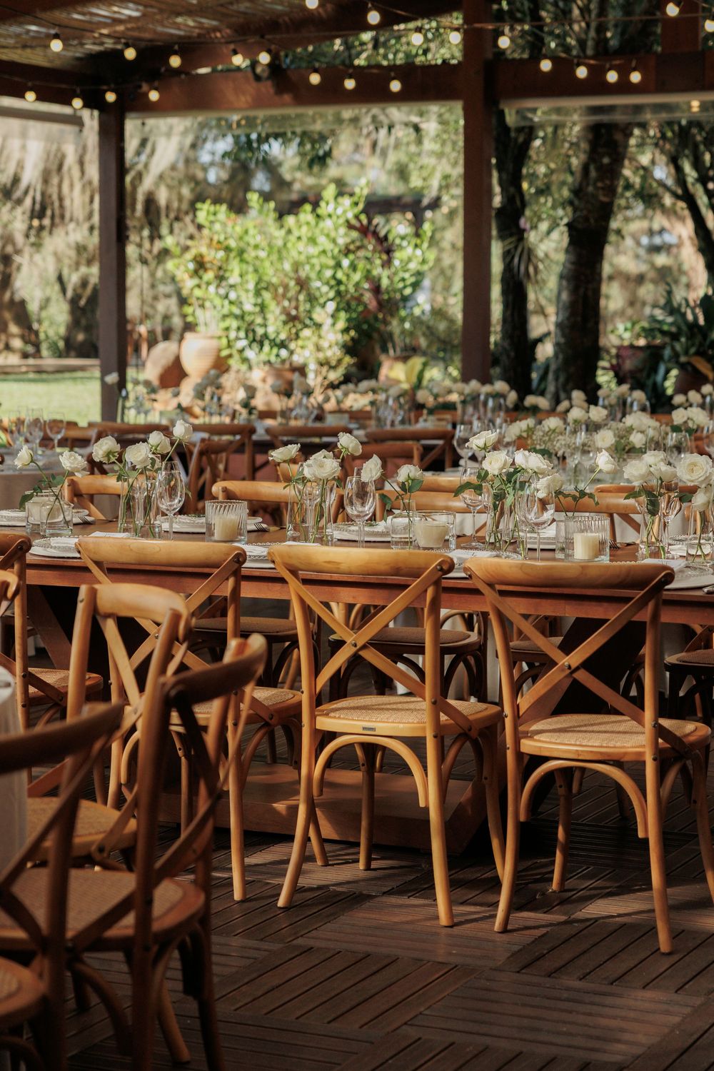Rustic outdoor wedding reception setup with wooden cross-back chairs and hanging string lights.