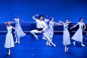 Ballet dancers in white dresses performing classical routine against blue backdrop.