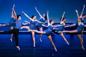 Dance ensemble in blue costumes performing synchronized leaps across stage.