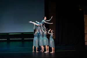Contemporary dancers in pale blue dresses creating lifting formation on dark stage.