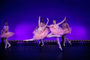 Ballet dancers in pink tutus perform synchronized routine under purple stage lights.