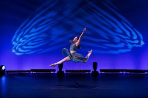 Dancer performs dramatic leap against blue illuminated backdrop on dark stage.
