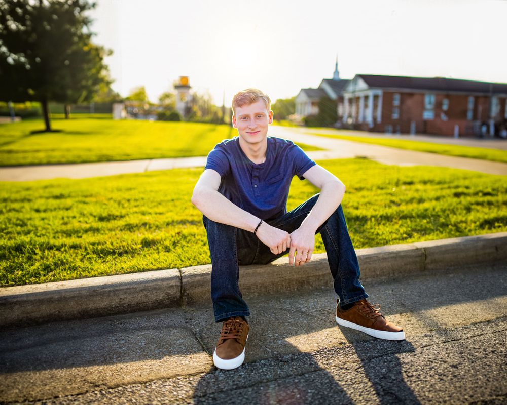 Person in blue shirt and jeans sits on curb during golden sunset with buildings in background.
