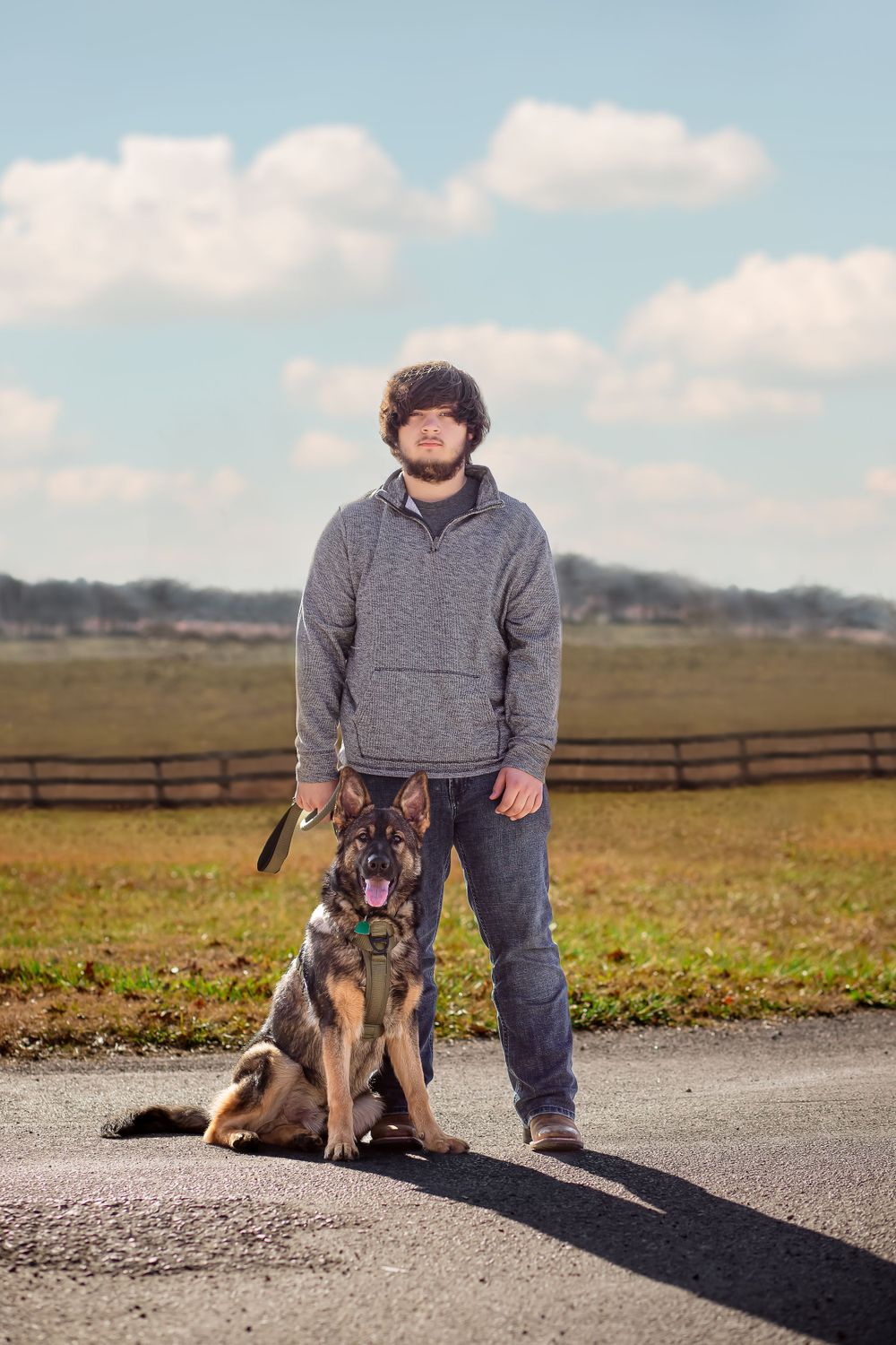 A German Shepherd stands at attention next to their owner in a rural field setting.