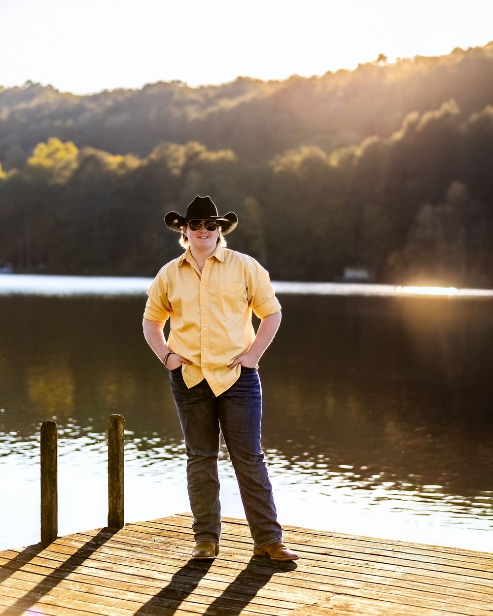 Person in a yellow shirt and cowboy hat stands on a wooden dock at sunset by a serene lake.