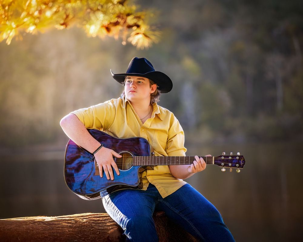 A person in a yellow shirt and blue jeans plays acoustic guitar while sitting outdoors under golden autumn leaves.