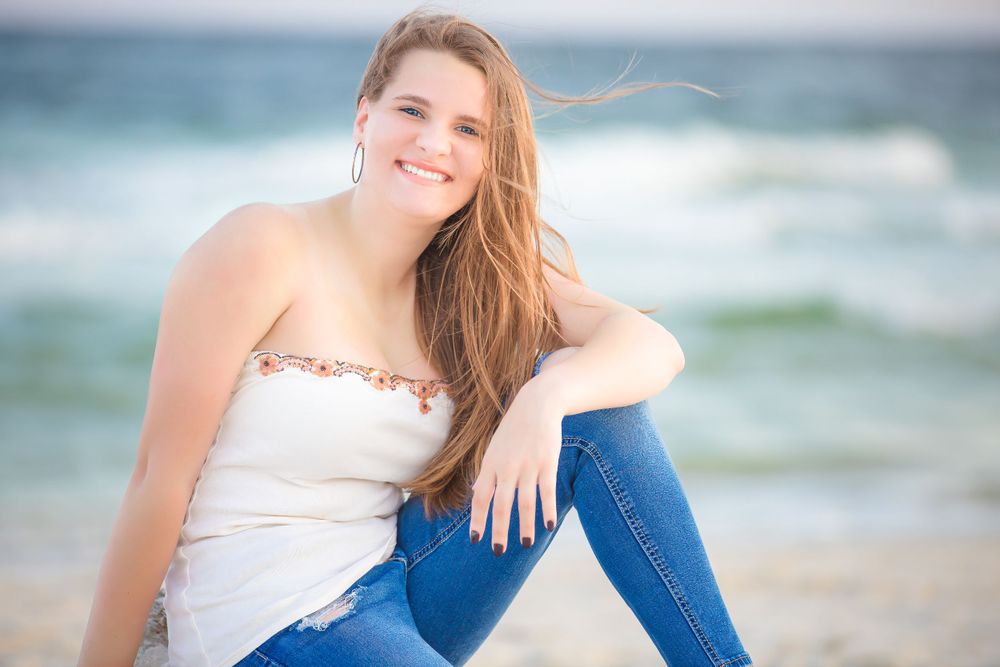 Portrait of a person in white top and jeans sitting casually on the beach with ocean waves in background.