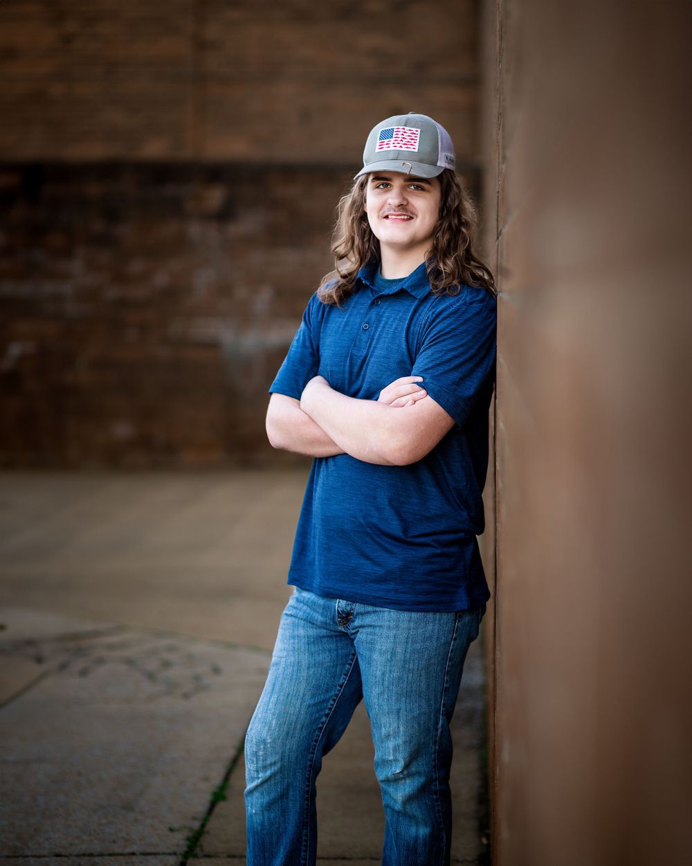 Person wearing baseball cap and blue polo stands with crossed arms against rustic wall.