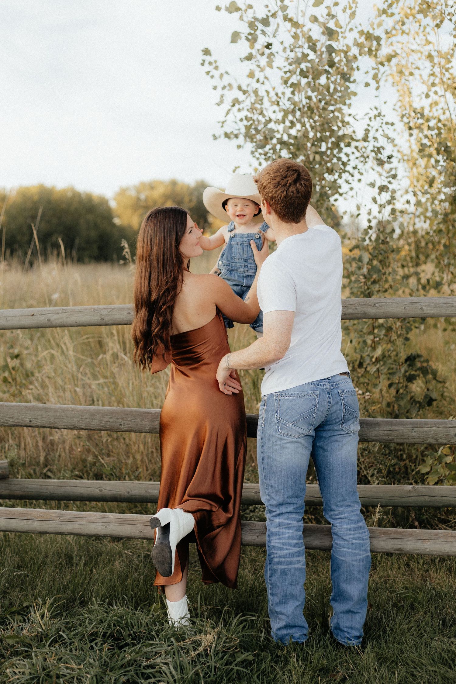 A young family stands by a rustic wooden fence at sunset in a golden field with trees in the background.