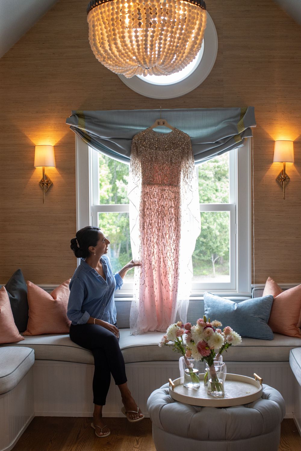 Bride admires her beaded pink wedding gown hanging in window of elegantly decorated bridal suite.