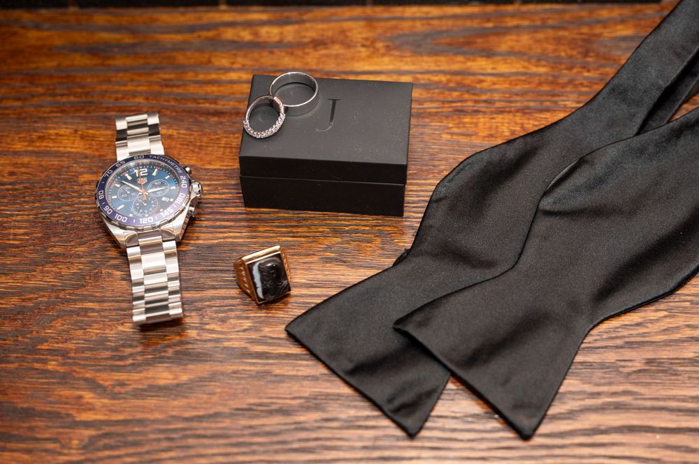 Silver watch, black ring box, cufflink, and black bow tie arranged on rustic wooden table for wedding day accessories.