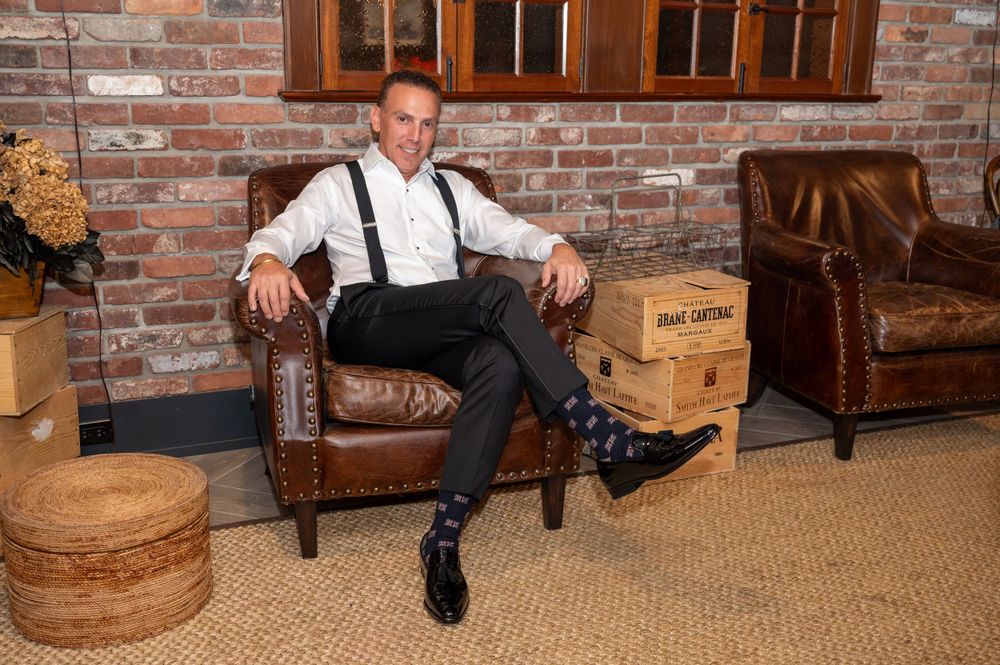 Groom relaxing in leather chair wearing suspenders and dress shirt in rustic brick-walled wedding venue.