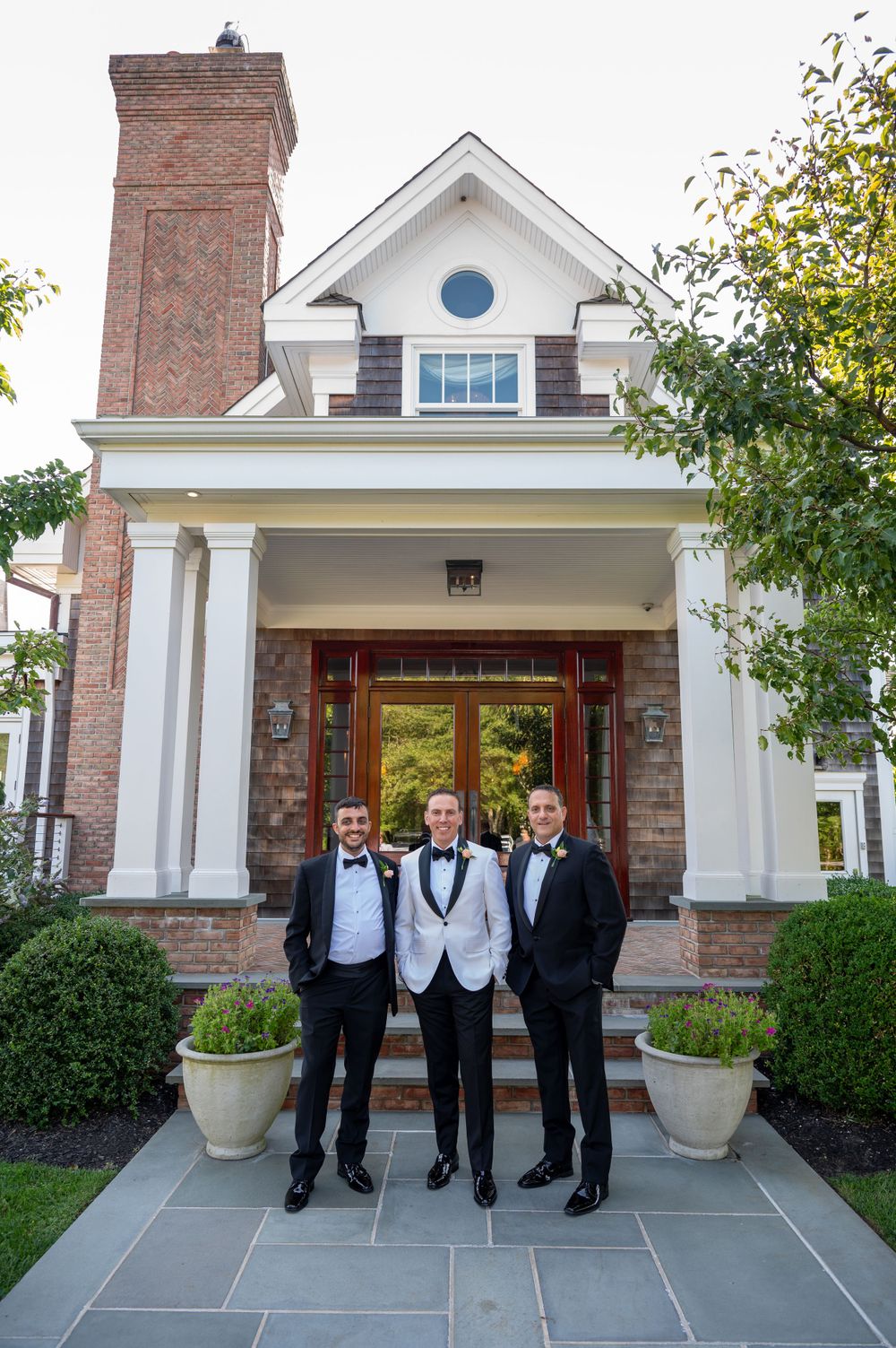 Groom in white tuxedo jacket stands with two groomsmen in black tuxedos outside elegant home.