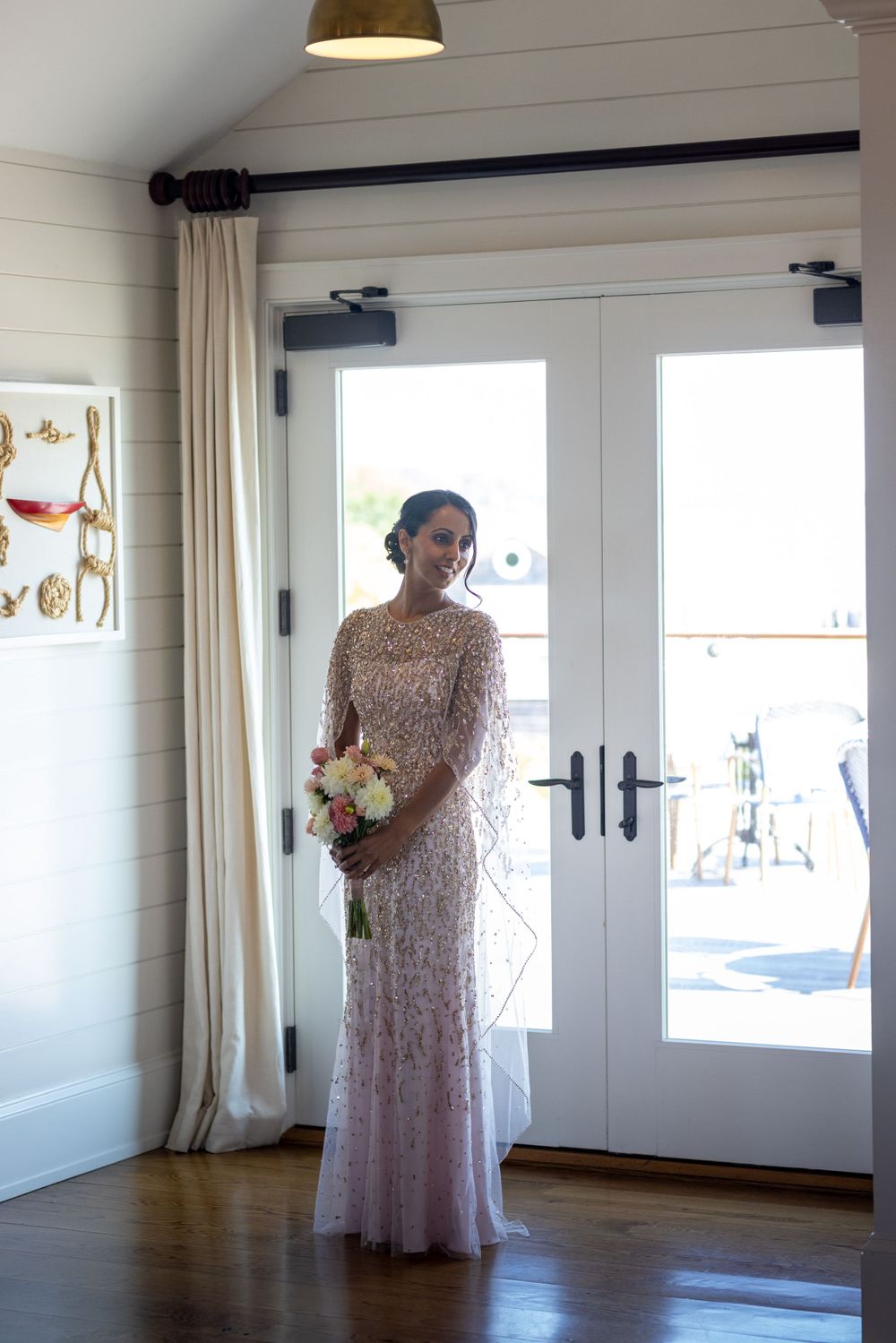 Bride in long-sleeve beaded blush gown holding bouquet stands before French doors in bright interior.
