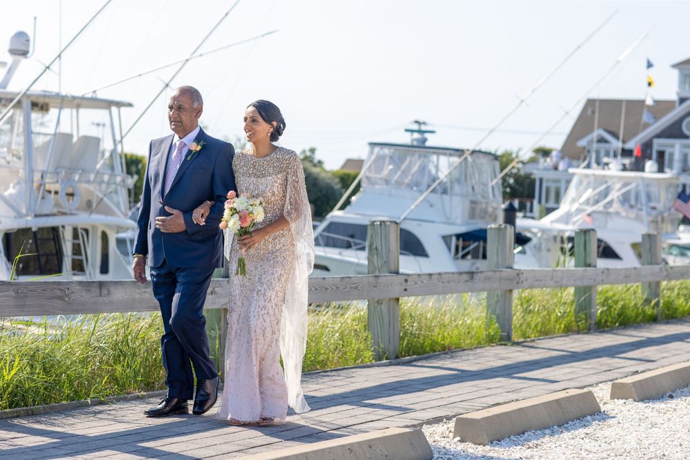 Bride in beaded gown and groom in navy suit walk along marina boardwalk with boats in background.