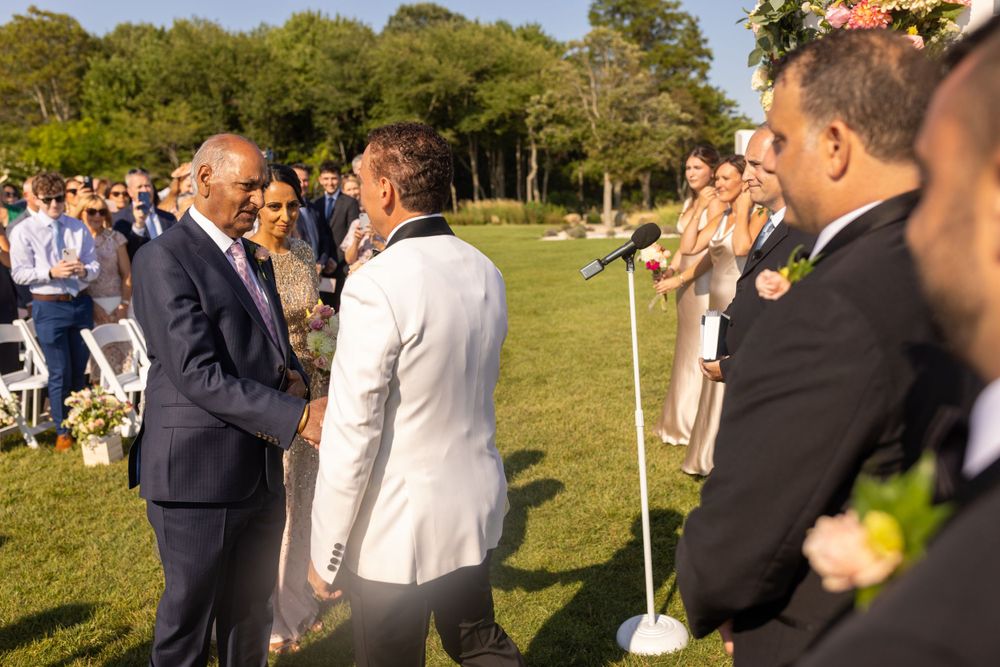 Father of bride shakes hands with groom in white jacket during outdoor wedding ceremony.