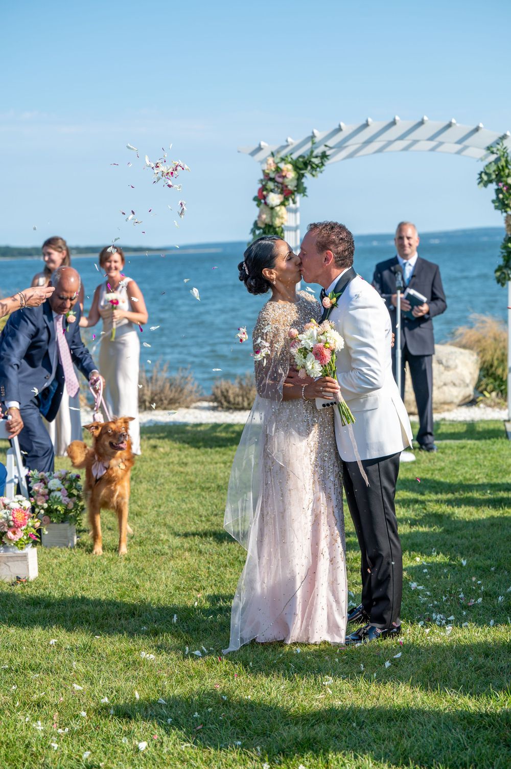 Bride and groom share first kiss at outdoor waterfront wedding ceremony as guests celebrate.