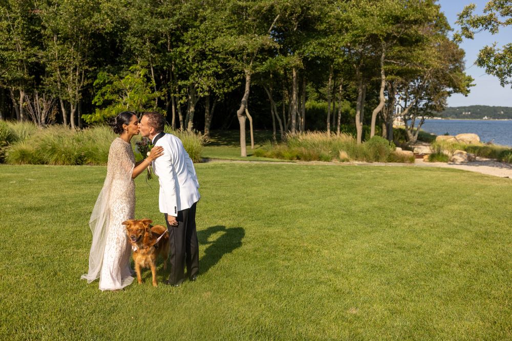 Bride and groom share a kiss on green lawn with their dog beside them at waterfront wedding venue.