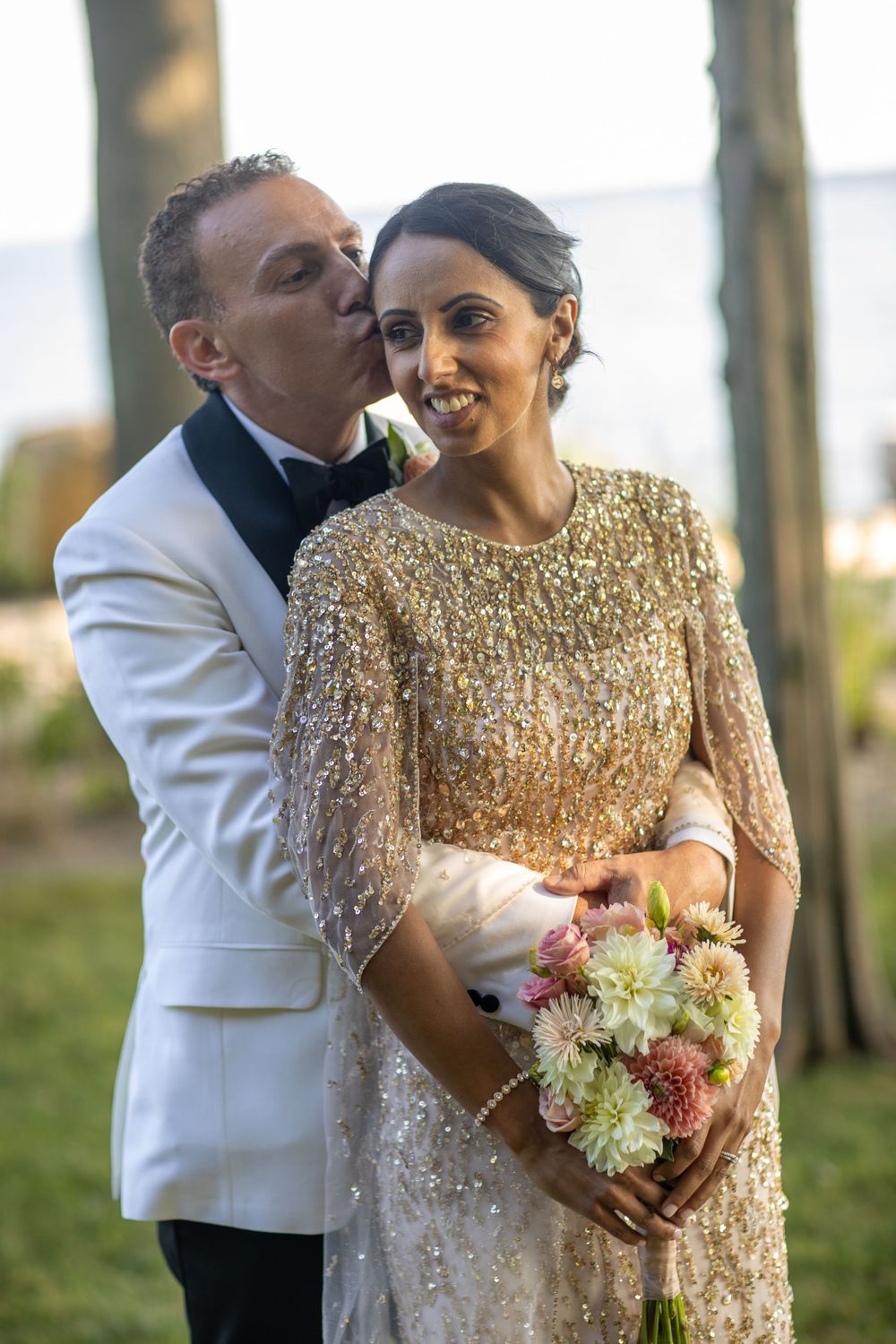 Groom in white tuxedo kisses bride wearing gold beaded gown while she holds floral bouquet.