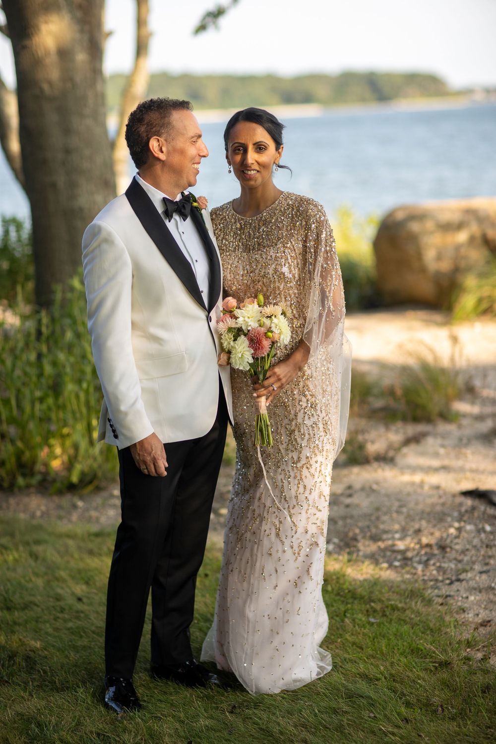 Bride in gold beaded gown and groom in white tuxedo jacket pose together near waterfront at outdoor wedding.