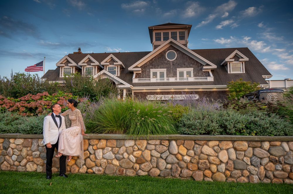 Newlyweds stand before Peconic Bay Yacht Club stone wall surrounded by colorful coastal gardens.