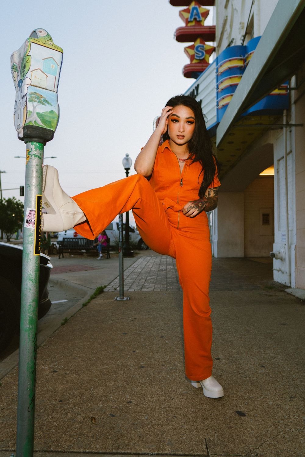 Model in bright orange jumpsuit strikes high-kick pose against urban storefront.
