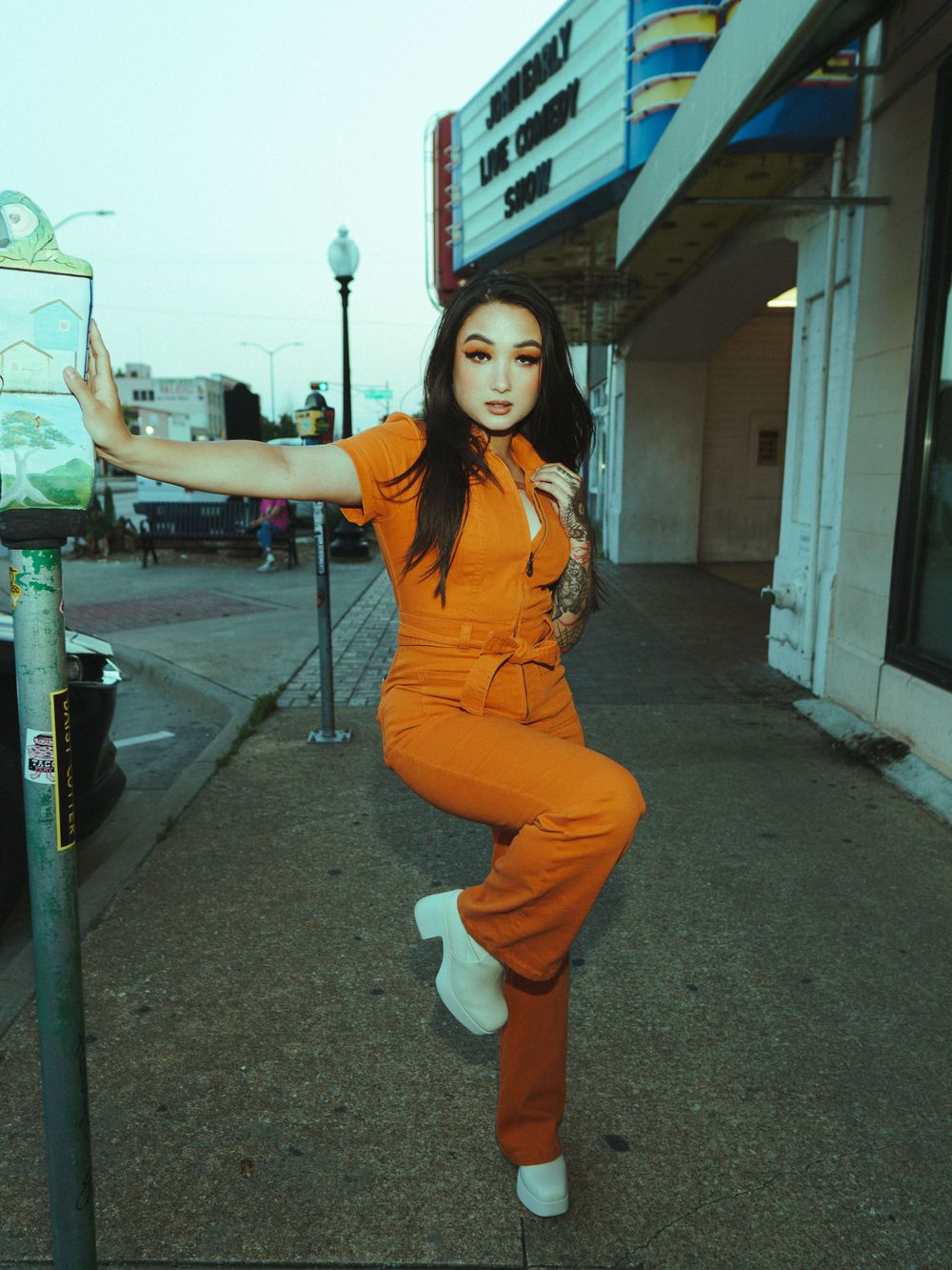 Woman in orange jumpsuit poses dynamically against retro storefront backdrop.