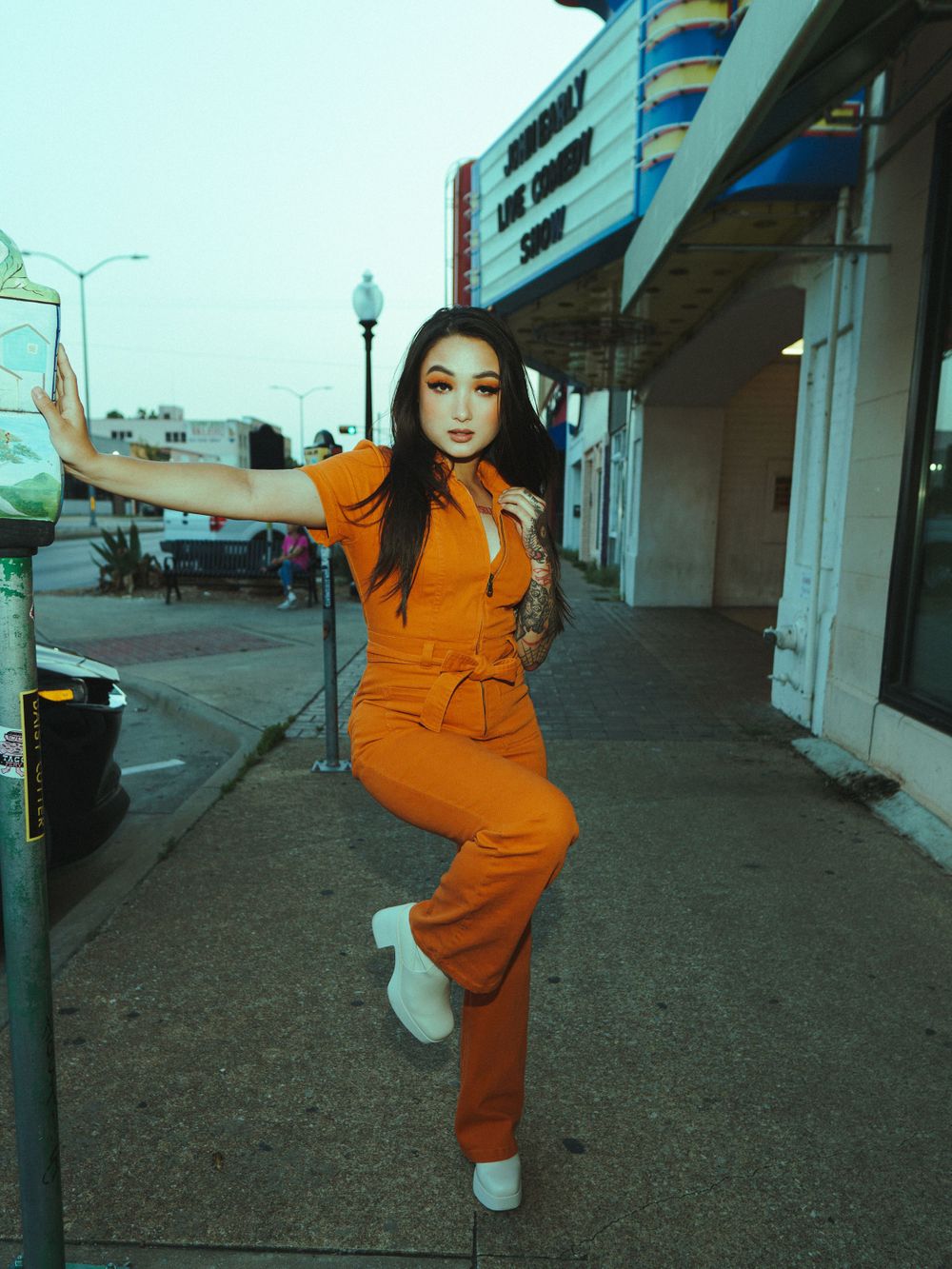 Model in vibrant orange jumpsuit poses elegantly against vintage storefront setting.